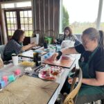Three women seated a table, all working on their lotus leaf paintings.