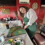 A woman standing and smiling at a table with a lotus leaf painting upon it.